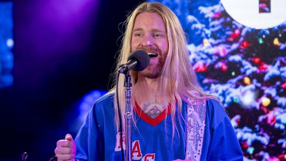 Sam Ryder in the BBC Radio 1 Live Lounge. Sam is a white man with long blonde hair, a short strawberry blonde beard and blue eyes. He wears a blue jumper with a red trim, just exposing the top of a chest tattoo and he has a sparkly guitar strap over his left shoulder. He sings into the microphone in front of him and is pictured in the BBC studio in front of a Christmas tree on a screen