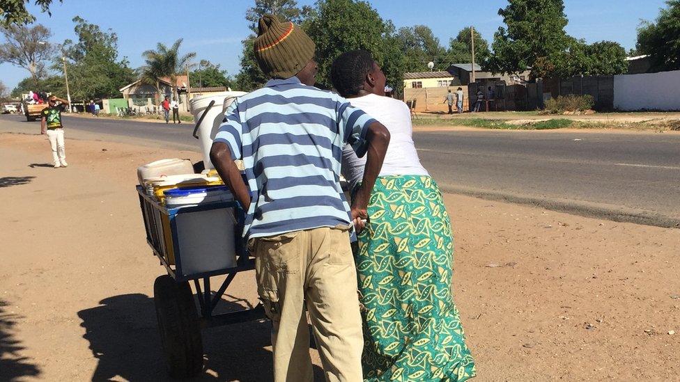 A man and woman push a wheelbarrow loaded with buckets of water.