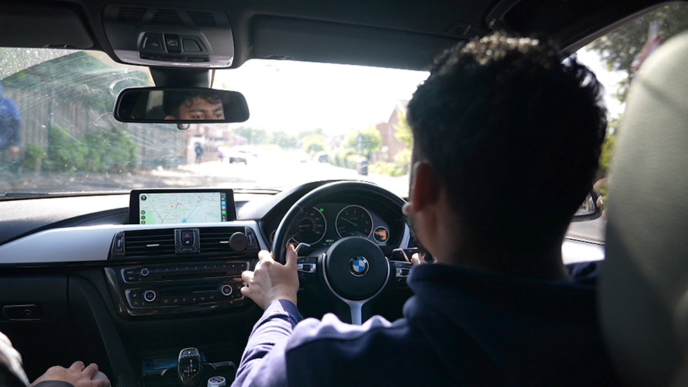 A man driving a car. The shot from the back seat towards the driver shows him looking forward over the steering wheel. You can see a glimpse of his face in the rear view mirror and a street scene out of focus through the windscreen