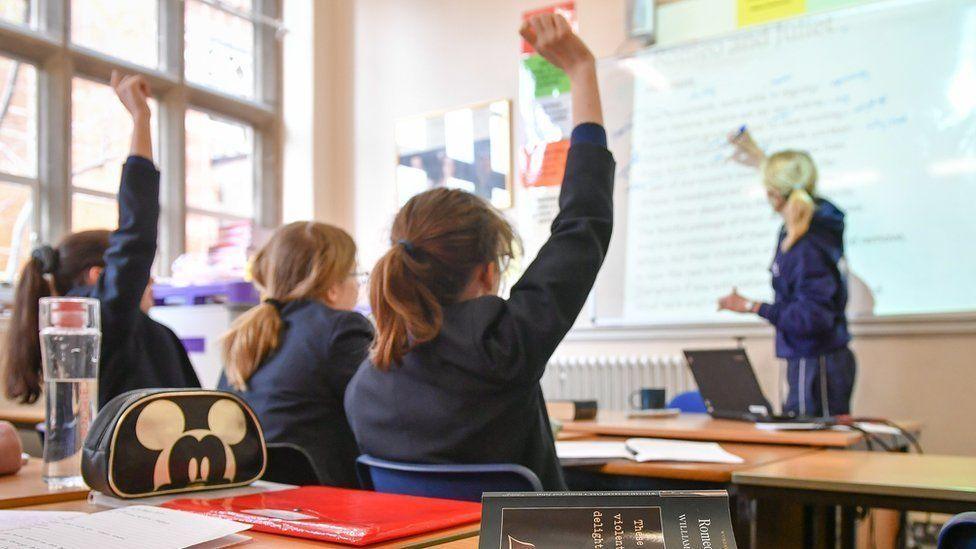 Children in an English lesson with their hands up and a Romeo and Juliet book open on the table