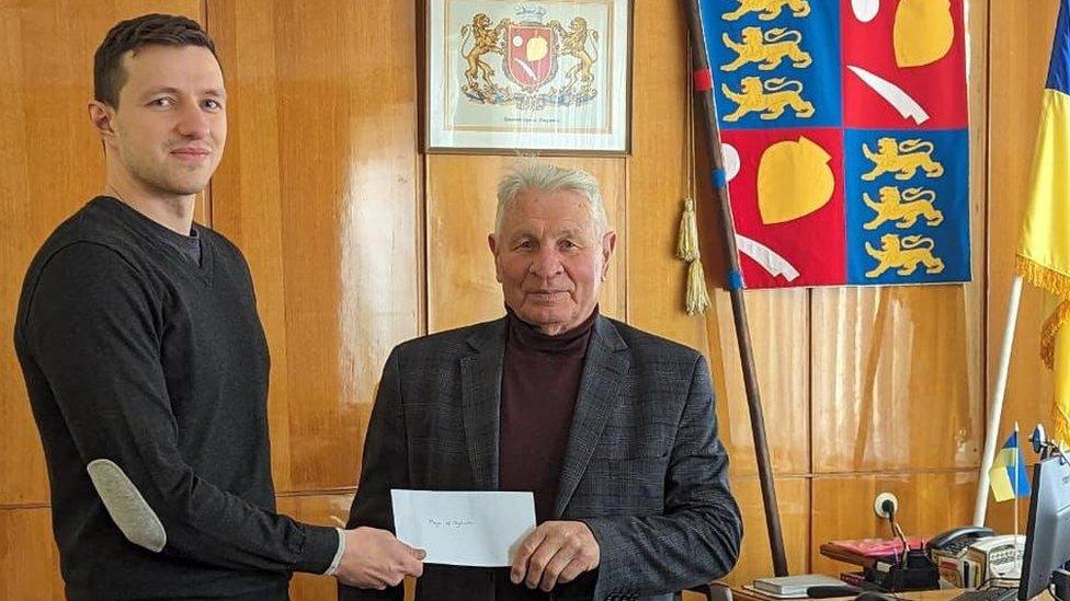 Young man with short dark hair presents older man with an envelope. A flag is visible behind them