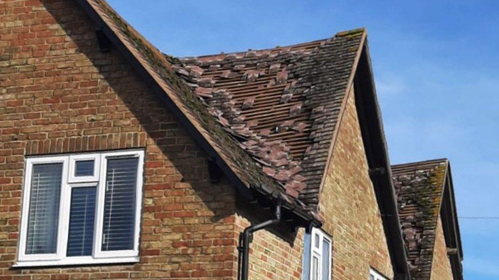 A red brick house. The roof and a few windows can be seen. The roof is missing lots of tiles from a storm and the foundations underneath are exposed.