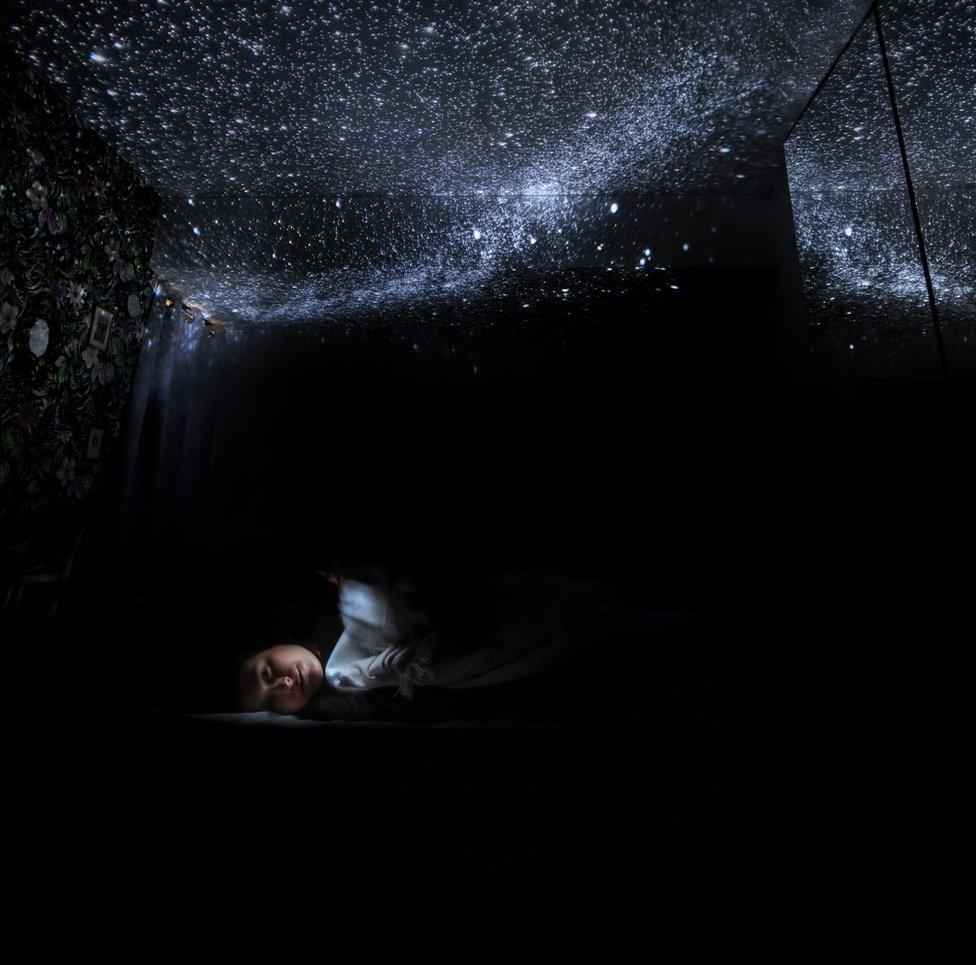 An image of a child in a bedroom with a constellation of lights above representing stars