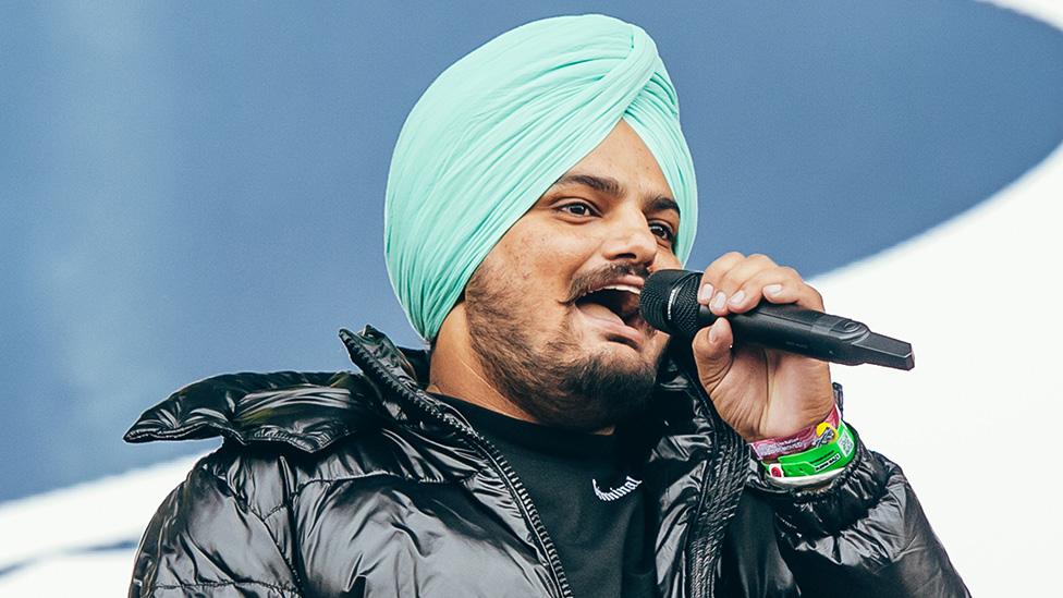 Sidhu Moose Wala wears a black puffa jacket and light blue turban as he performs at Wireless Festival in 2021. He's holding the microphone to his mouth and appears to be mid-rap.