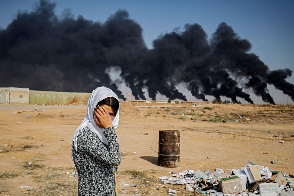 A woman covers her face against a background of smoke plumes