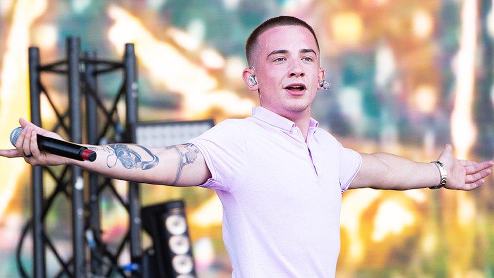 ArrDee looking at the crowd with his arms spread out with a mic in his right hand. he's wearing a light pink tshirt with a collar. He has an ear piece in his ears. Behind him is staging with multi coloured lights and metal railing holding up light and sound equipment.