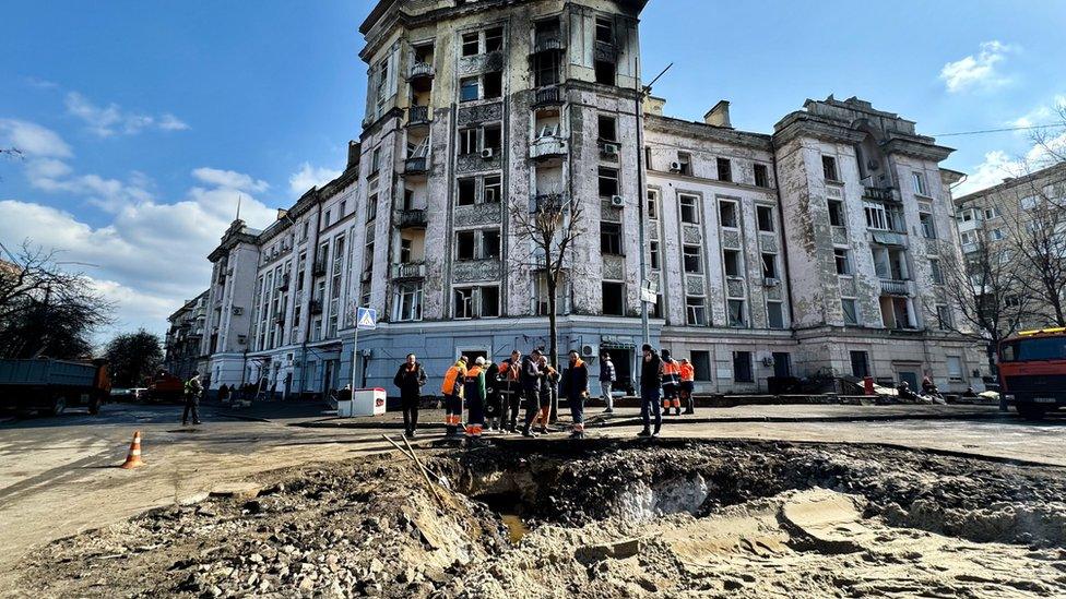 A large crater in the ground in front of an apartment building with fire damage at the windows