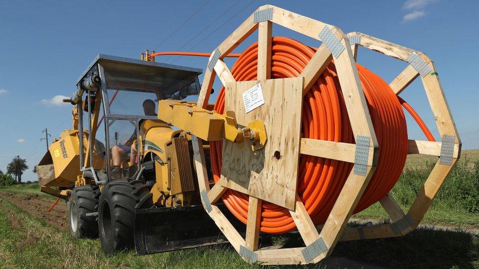 A large work vehicle carrying a mammoth spool of reel rides across open green grassland