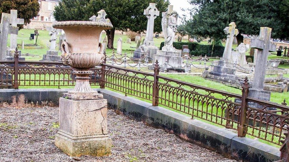 Urn to mark a grave in a cemetery showing cracks in the stonework, surrounded by brown railings