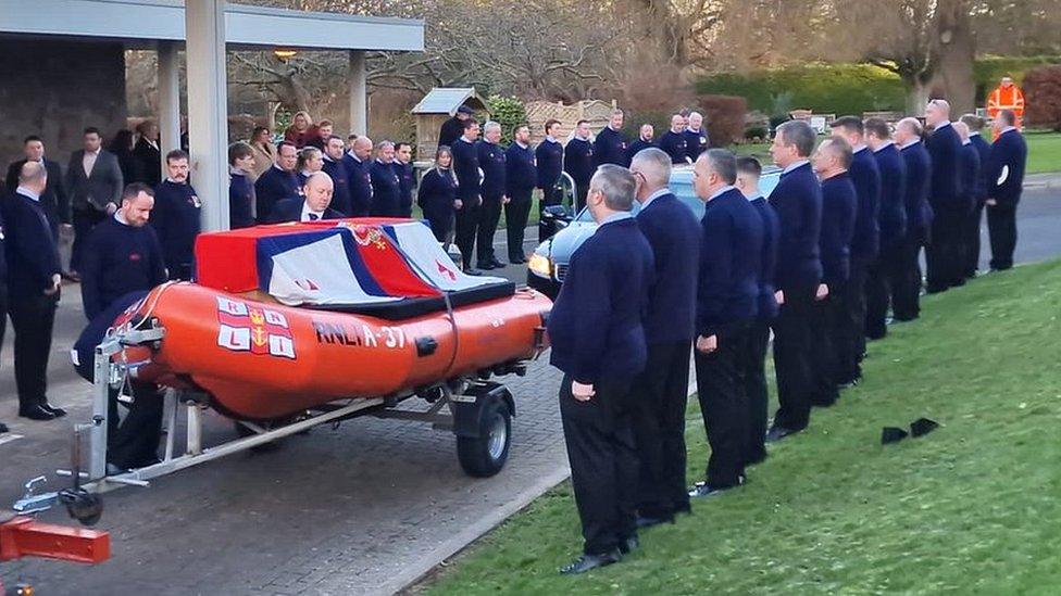 People in dark clothes lining the path while a lifeboat with a coffin on top is driven past