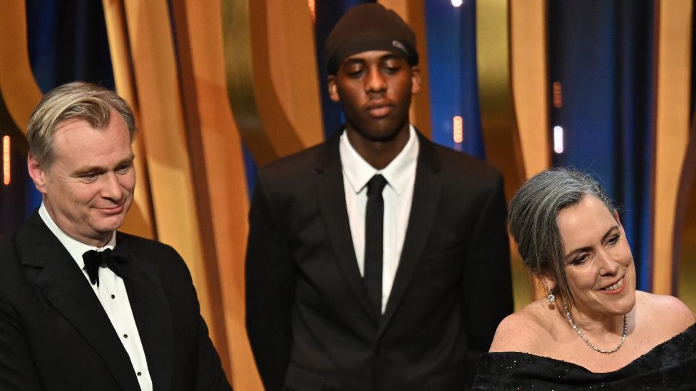 Cillian Murphy, Christopher Nolan, and Emma Thomas accept the Best Film Award for 'Oppenheimer' on stage during the EE BAFTA Film Awards 2024 at The Royal Festival Hall on February 18, 2024 in London, England, while a YouTube prankster stands in the background