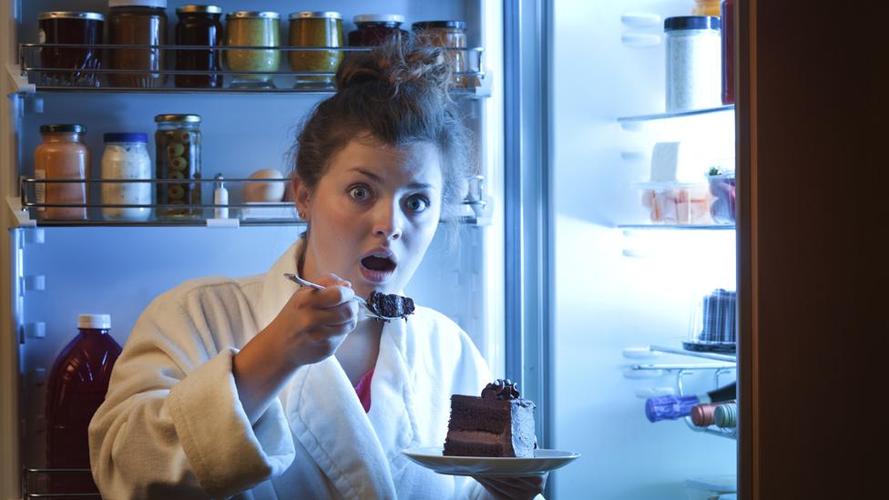 Woman in dressing gown tucking into chocolate cake in front of fridge