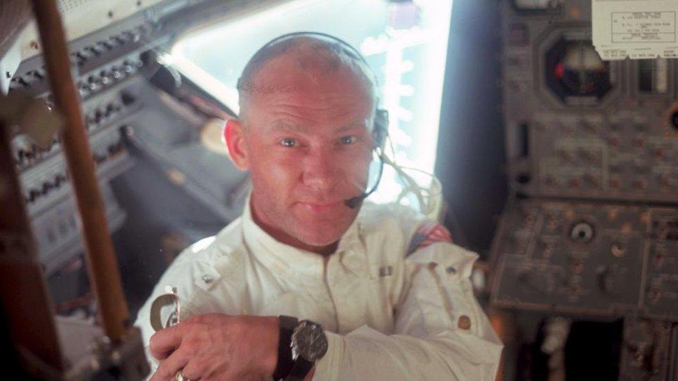 Buzz Aldrin wearing a spacesuit and a helmet with a headset inside the Apollo 11 lunar module, surrounded by knobs and buttons