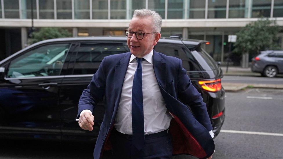 Michael Gove wearing a navy suit. The red trim of the jacket is just visible as it gets slightly blown in the wind. He is looking off to the right, with a car behind him.