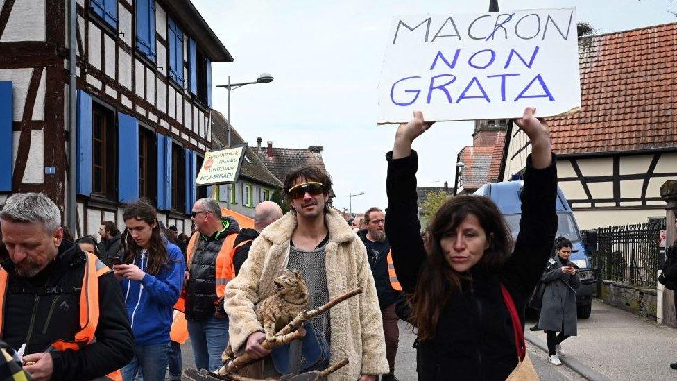 A demonstrator raises a placard during a protest against a visit of French President Emmanuel Macron to Mathis, a company specialised in large wooden buildings, in Muttersholtz, eastern France on April 19, 2023