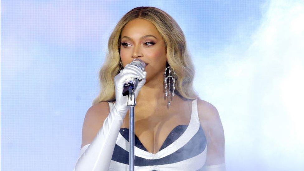 Beyoncé performs onstage during the "RENAISSANCE WORLD TOUR" in New Jersey. She is wearing a black and white striped dress and ha a white full arm length glove on right hand which is holding the microphone in front of her mouth. She has long blonde hair and has a large diamond necklace in her left ear. Behind her the background is light blue with white smoke visible to her right.