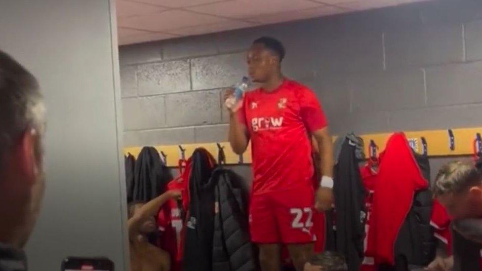 A young black man in a red sports kit singing a song using a water bottle as a mic