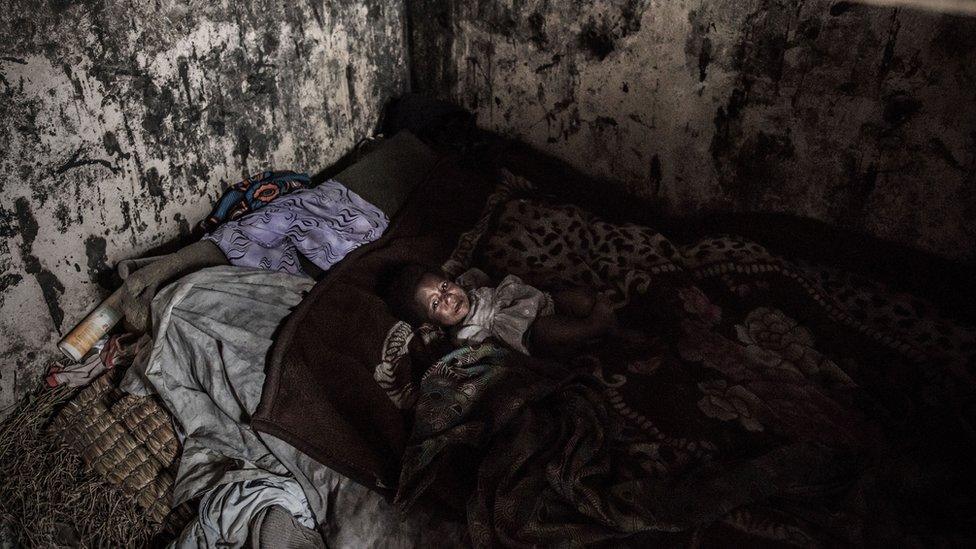 young boy looking drawn and tired lies in a dark, damp-looking room inside a thatched hat