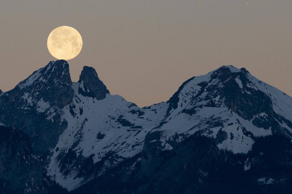 The full moon sets behind the mountains including the twin peaks of Les Jumelles in the Chablais Valaisan, seen from Fenalet-sur-Bex, Switzerland, on 18 January 2022