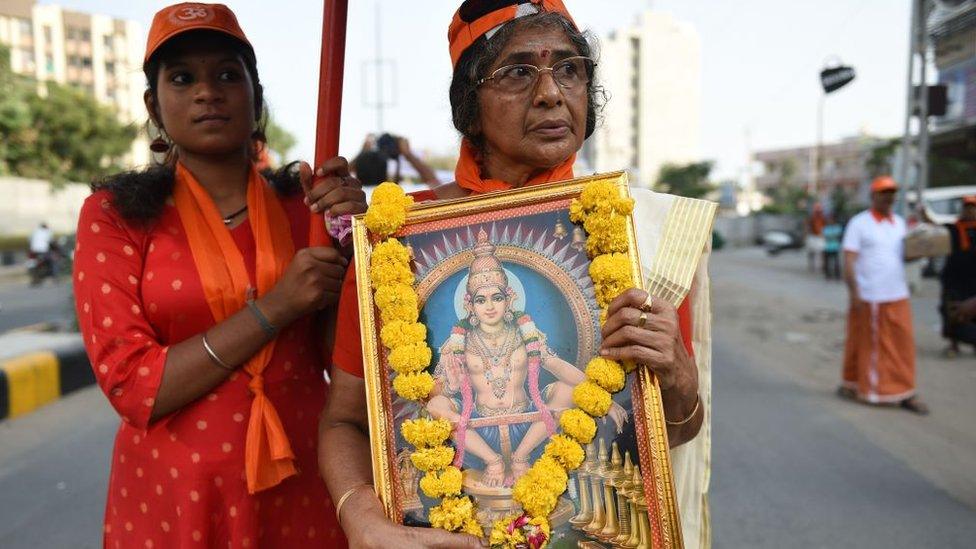 A woman holding a framed poster of a Hindu god followed by another woman