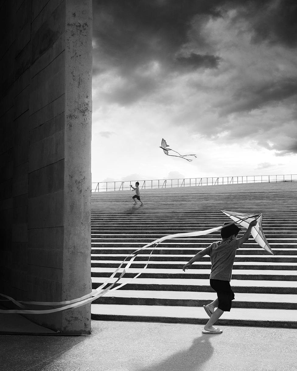 A black and white photo showing two children playing with kites on the steps of Teopanzolco Cultural Center in Mexico with dark clouds in the sky