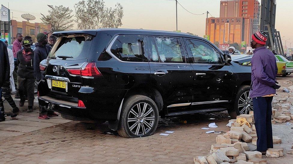 Locals congregate around a black SUV vehicle with holes in the metal on the side