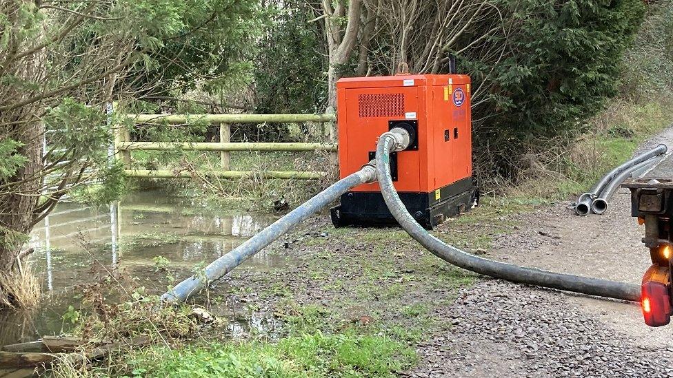 A large orange box-like pumping machine next to a waterlogged area with large pipes coming from it