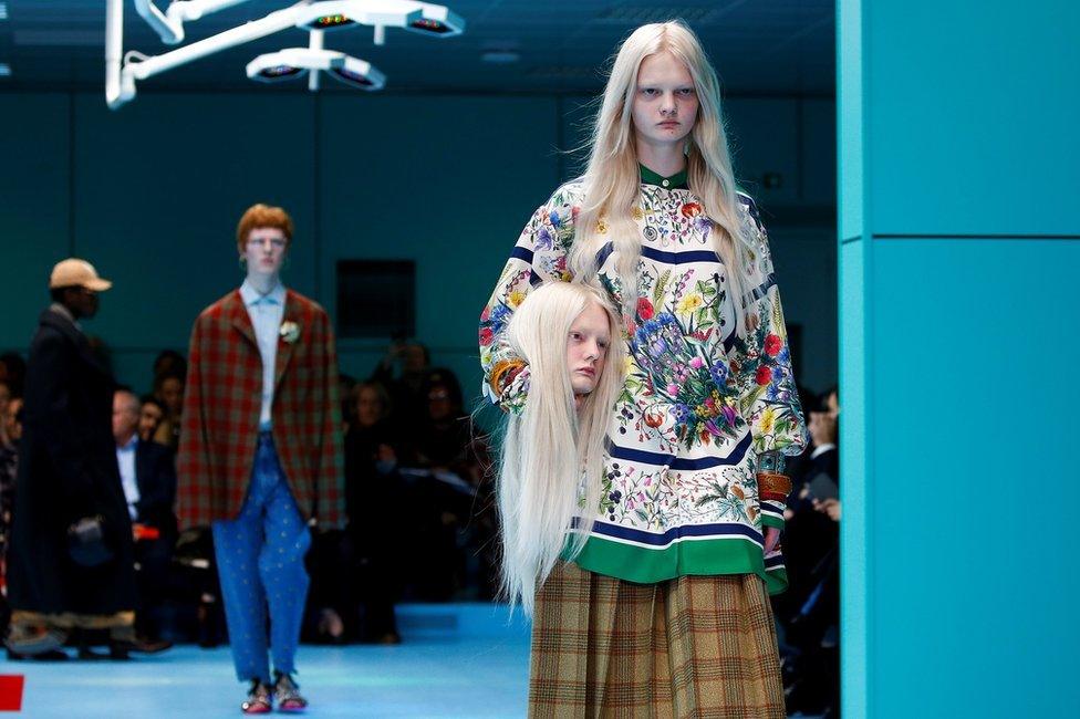 Female model in floral dress walks down the catwalk holding a mannequin head that is a replica of her head