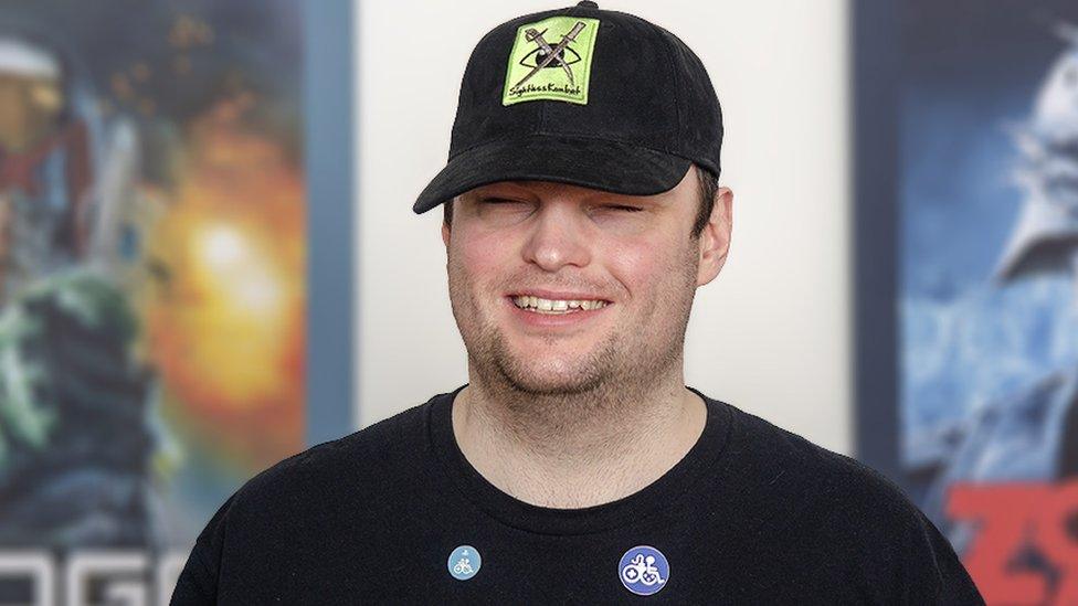 A young man smiling. The logo on his black baseball cap is a lime green square with a pair of crossed swords - a curved, katana-style blade and more elaborate, serrated weapon - over the top of an eyeball graphic. Underneath the words "Sightless Kombat" are written in black script. He's wearing a black t-shirt with an RNIB (Royal National Institute of Blind People) logo on it, and two round pin badges just below the neck line. Both of them have a white, graphic design that mixes a gaming joypad with the seated stick figure more commonly seen with a wheelchair on disabled signage. Behind him two pictures hang on the wall - one shows comic book character Judge Dredd and the other a zombie soldier whose eyes glow yellow underneath his tattered military helmet.