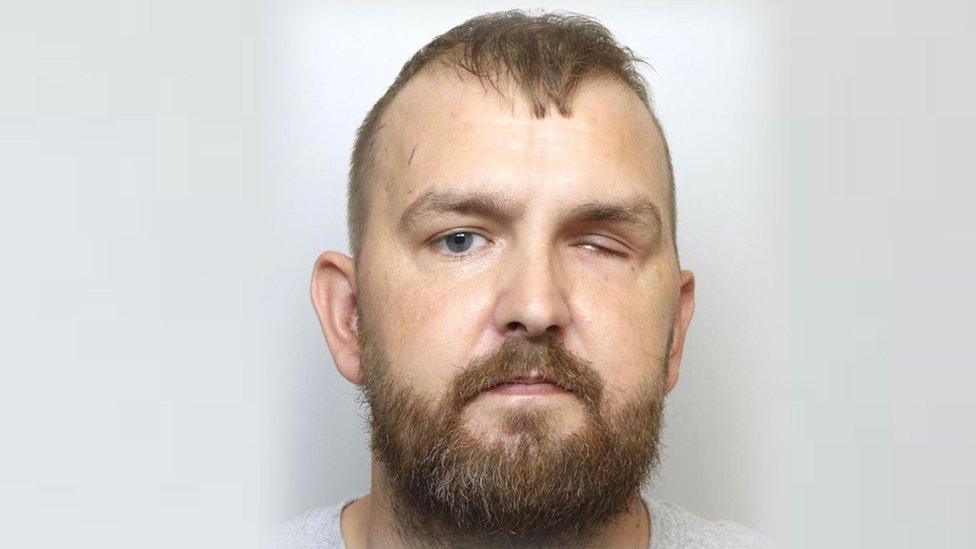 Man with short brown hair and beard in police 'mug' shot. One eye is almost entirely closed.