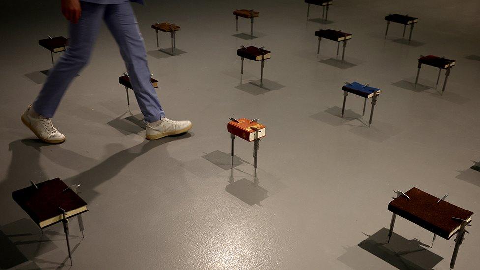 A gallery worker walks through an installation by Rwandan artist Francis Offman showing books including a copy of the bible belonging to his mother held up by callipers