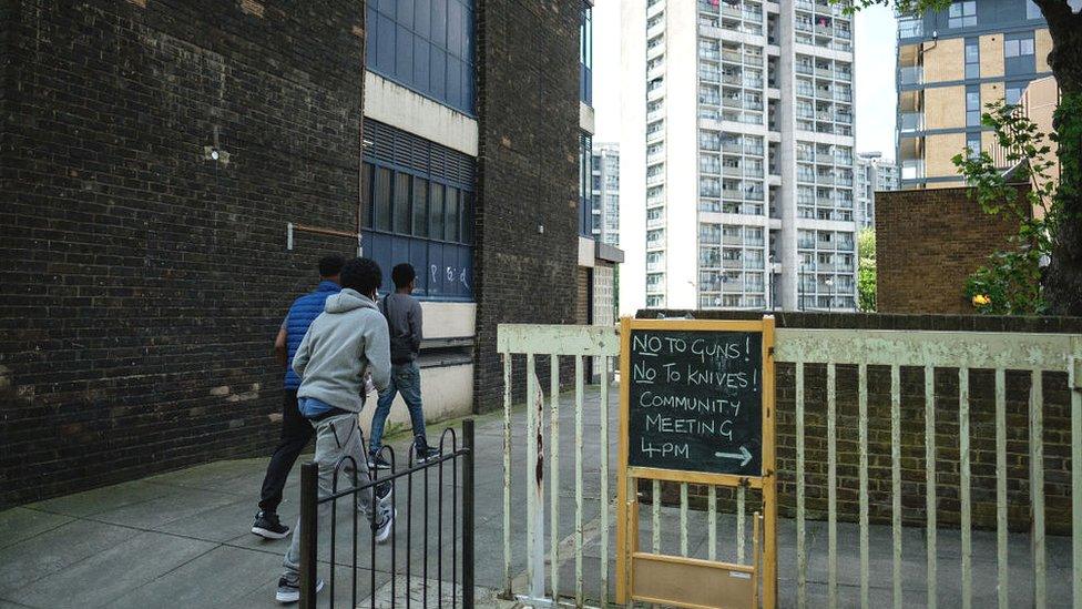 3 boys walking on housing estate near sign reading no to guns! no to knives!