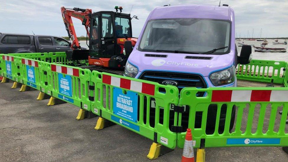 Blue CityFibre van and a yellow digger are parked behind green railings