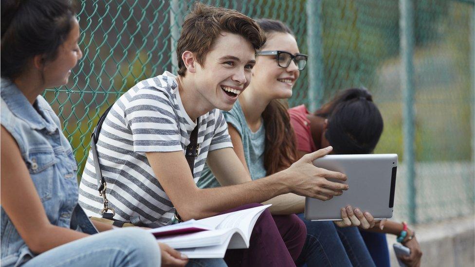 Friends laughing together at school, holding tablet and books