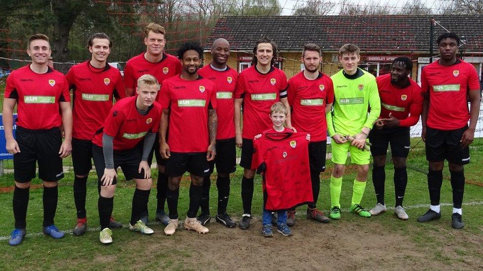 Liam standing amongst eleven footballers. He is holding a signed shirt.