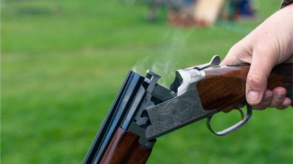 A close up of a person holding a shotgun with smoke coming out of the barrel with grass in the background.