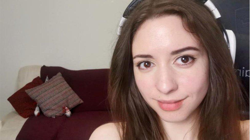 A picture of Anita smiling at the camera, in front of a blank wall with a sofa in the middle ground.