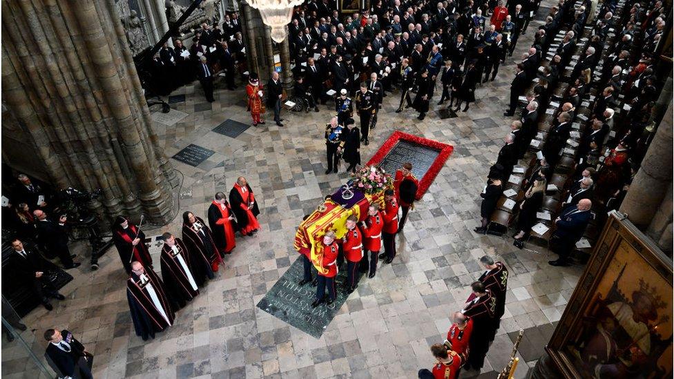 a still from above shows the coffin in westminster abbey held by pall bearers with funeral guests dressed in black seated nearby