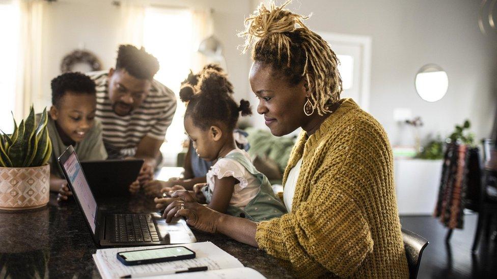 A mother working from home while holding a toddler, with her family by her side