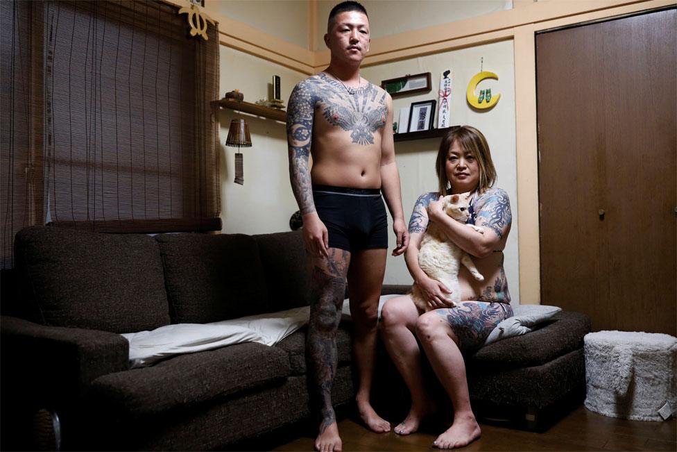 A tattooed woman and her adult son pose in their home