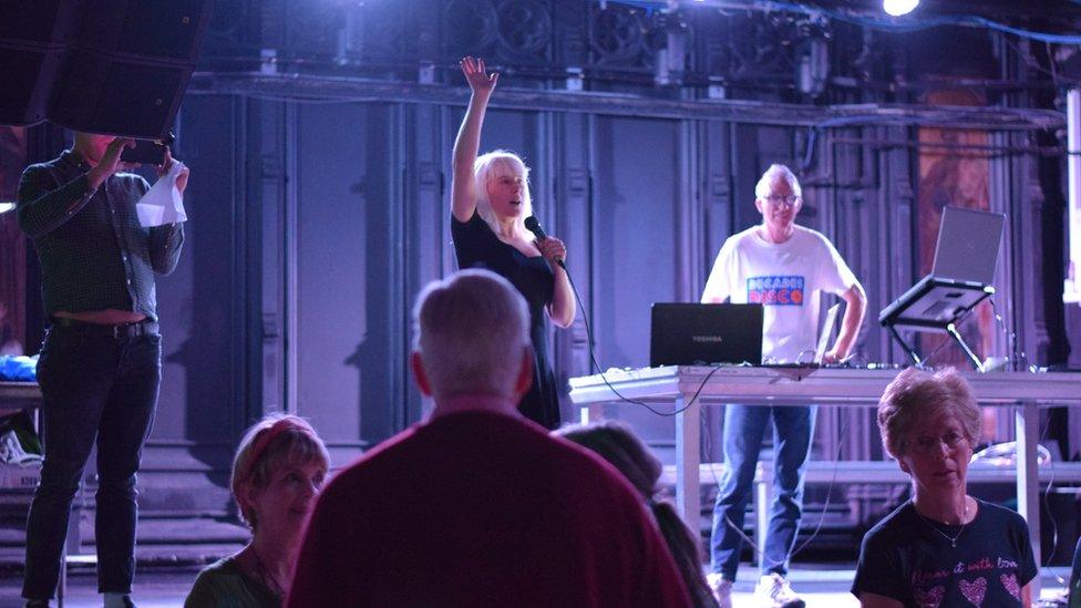 A blonde woman is holding a microphone with her hand in the air. She is standing next to the DJ and in front of an audience