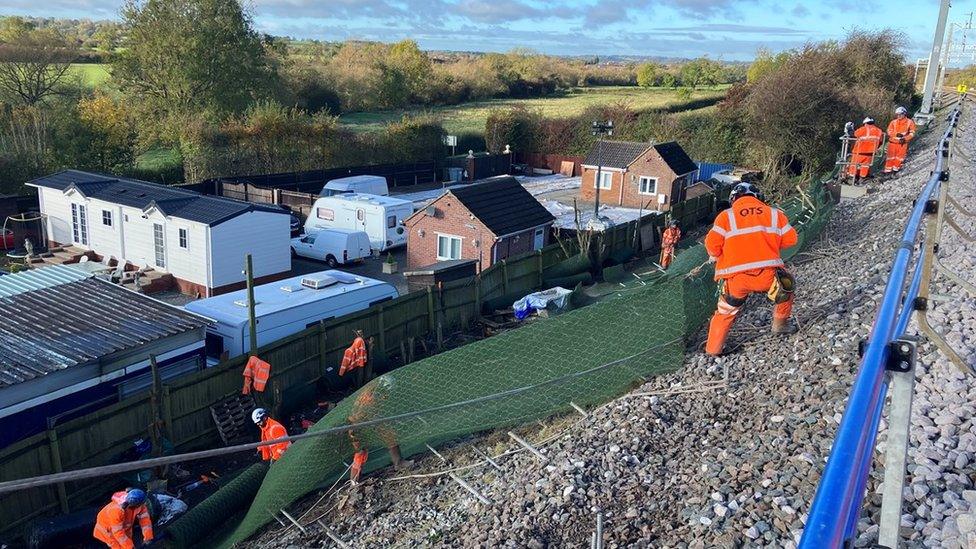 Workers in orange hi-viz erecting some green netting alongside a railway line