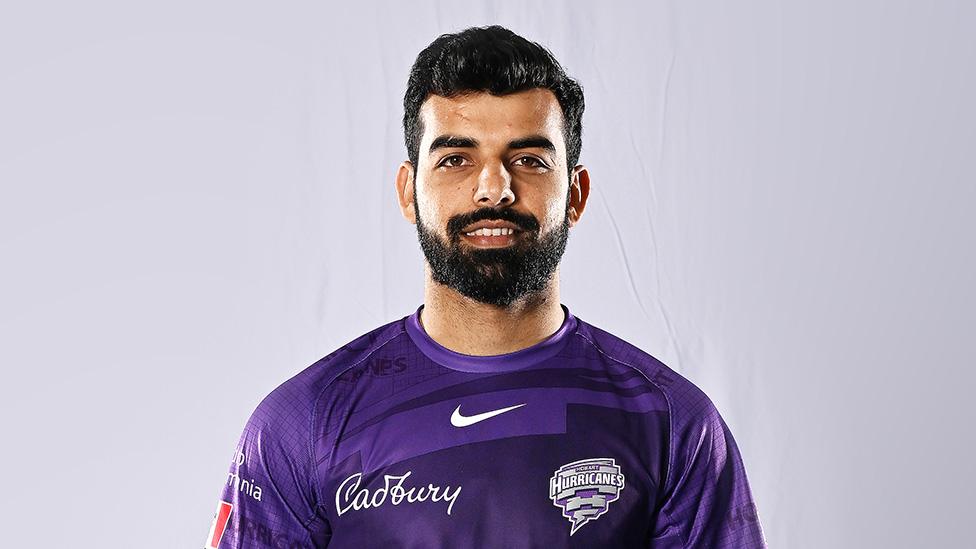 Shadab Khan against a white studio background. He's got short hair and a trimmed but thick-looking dark beard. He's smiling and wearing a purple t-shirt with a Nike "swoosh" logo over the upper chest. Below that on the camera left is an embroidered Cadbury logo - in cursive writing and white text. On the right is the Hobart Hurricanes' team logo - the word Hurricanes in angular white text over a grey, precisely-drawn shaped made up of curved rectangles that decrease in size from top to bottom to create the effect of a hurricane or funnel cloud.