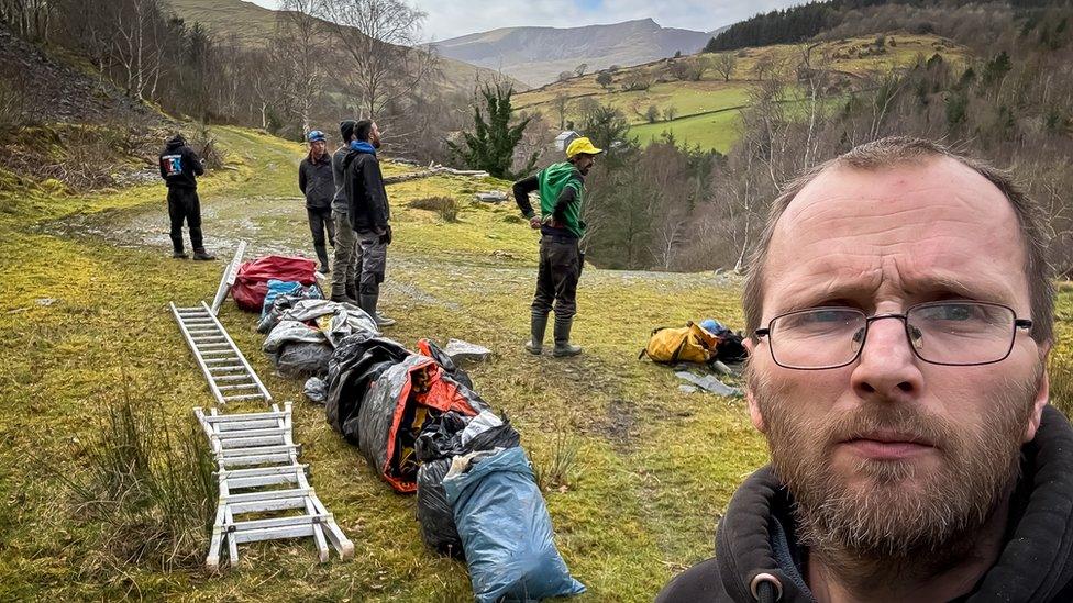 Gareth Jones selfie with all the rubbish and ladders on grass outside the mine, with the volunteers looking out across a Welsh valley