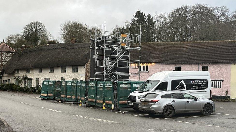 Scaffolding and construction fencing round an area in a quaint village with thatched cottages