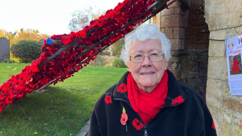 Woman with white hair and glasses, wearing a black coat, a red scarf and poppy stands beside a line of poppies