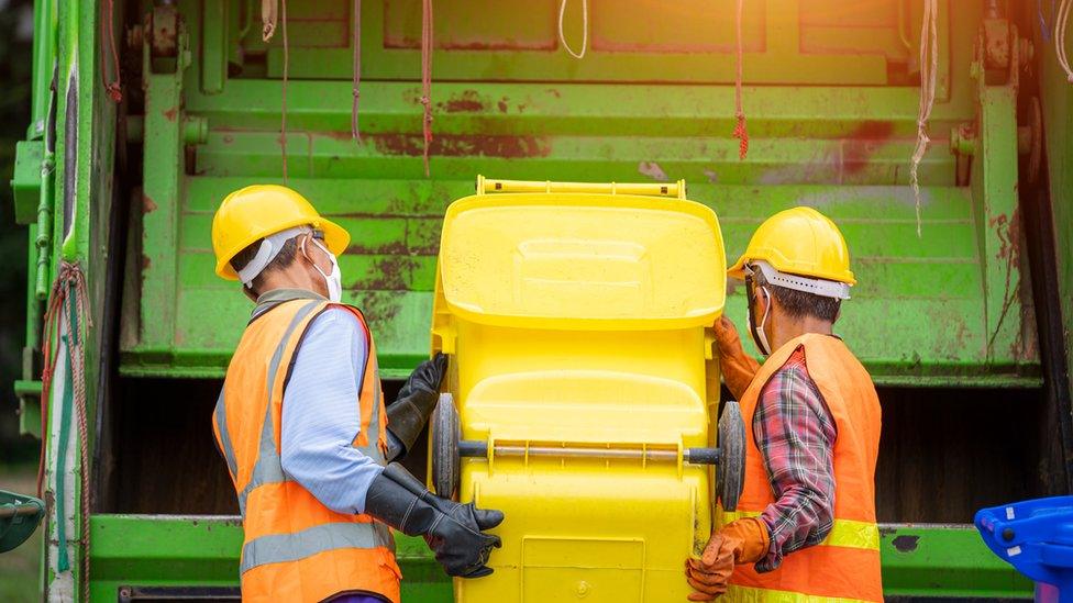 Two refuge collectors load waste into the back of a green bin lorry from a bright yellow wheelie bin