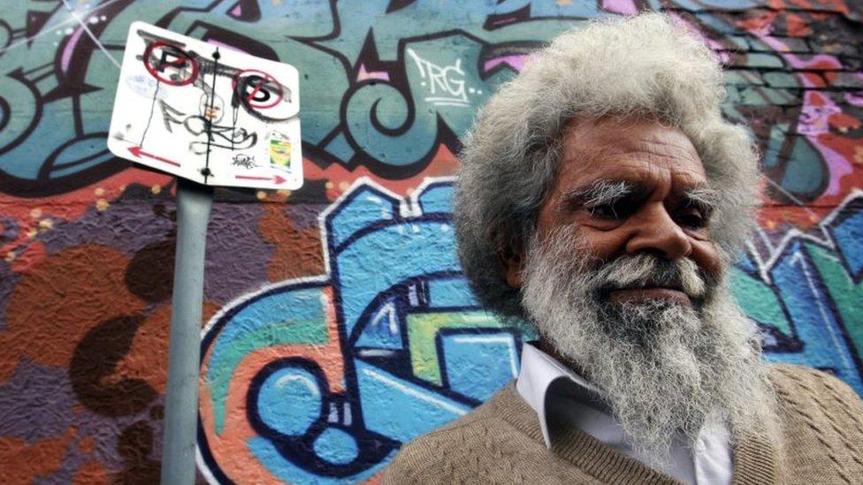 A man with white hair stands in front of a wall covered in graffiti