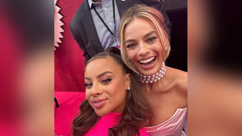 Danica Taylor and Margot Robbie posing for a selfie at the Barbier premiere, Margot looks very happy and Danica is smiling. Margot is wearing a pearl necklace and has her blonde hair tied up while Danica has her brunette hair worn down. Both are wearing pink dresses