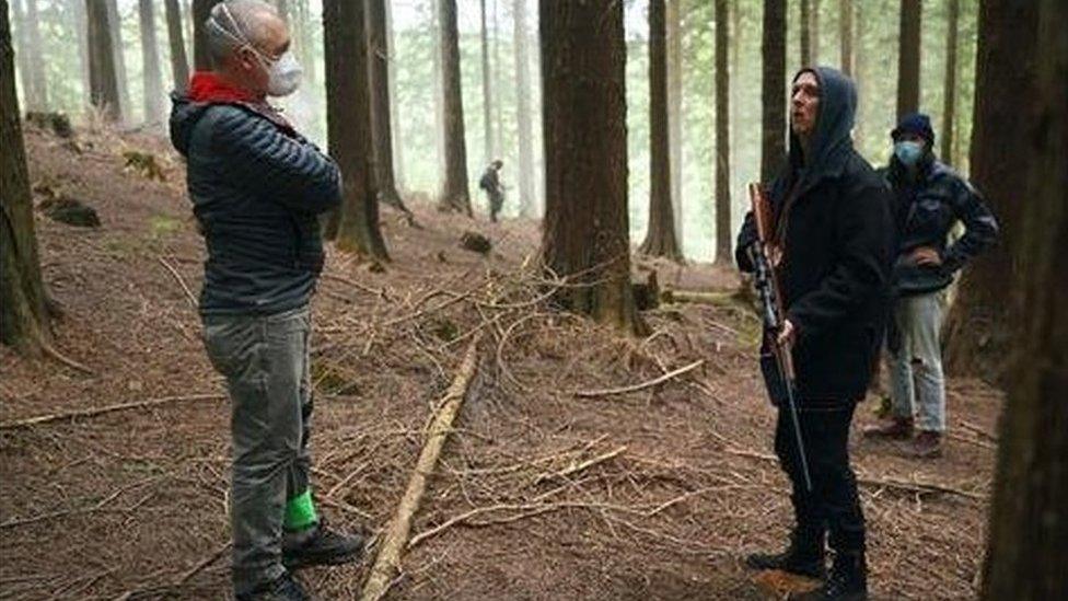 A crew member wears a mask, while an actor holds a fake rifle, surrounded by forest